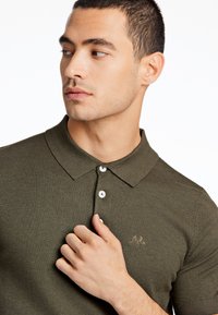 Olivgrön stickad piké med krage, tvåknappsstängning och subtil broderad logotyp på bröstet. Slät tygtextur.