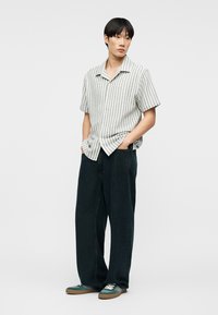 Νέος άνδρας που στέκεται με τα χέρια στις τσέπες, φοράει κοντομάνικο ριγέ πουκάμισο με κουμπιά, φαρδιά μαύρη παντελόνια και πράσινα αθλητικά παπούτσια.