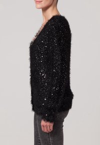 Svart plyschtröja med paljetter, V-ringad design, långa ärmar, strukturerad tyg. Matcher med grå denim för kontrast.