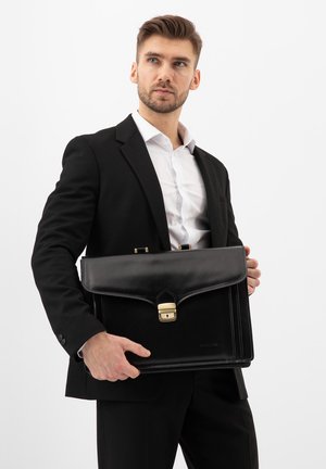 Muž v černém obleku a bílé košili držící černou koženou aktovku se zlatou sponou, mírně se dívá stranou.