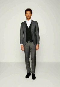 Costum gri în carouri, cu o croială ajustată, dotat cu o vestă cu trei butoane, cămașă albă și pantofi negri. Material neted și design structurat.