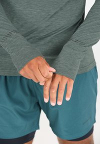 Langarm, dunkelgrünes Sportoberteil mit Daumenlöchern und strukturierten Bündchen, kombiniert mit türkisfarbenen Shorts. Aus glattem, feuchtigkeitsableitendem Material.
