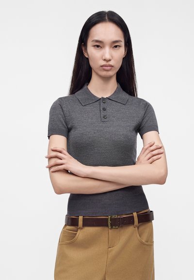 Jeune femme aux longs cheveux foncés portant un polo gris à manches courtes et un pantalon beige, debout, les bras croisés, devant un fond blanc.