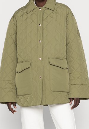 Olivgrüne gesteppte Jacke mit großen Fronttaschen und goldenen Druckknöpfen, getragen über einer weißen Hose.