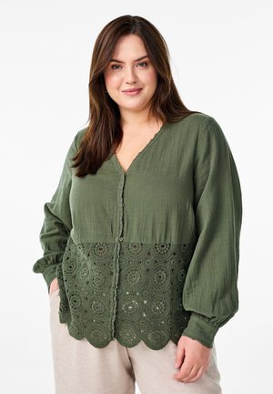 Blouse verte avec un décolleté en V, fermeture à boutons et manches longues. La moitié inférieure présente un design en dentelle au crochet à bord festonné avec des motifs circulaires.