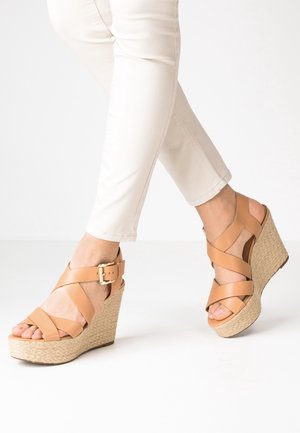 Fødder iført beige læder kilehælssandaler med krydsede remme og guldlås, kombineret med hvide, slim-fit bukser mod en ensfarvet baggrund.