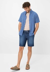 Camicia a maniche corte blu e bianca a righe, t-shirt beige sotto, abbinata a pantaloni shorts in denim scuro e sandali marroni. Outfit estivo casual.