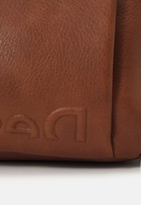 Surface en cuir brun texturé avec un logo partiellement embossé en bas à gauche sur fond blanc.