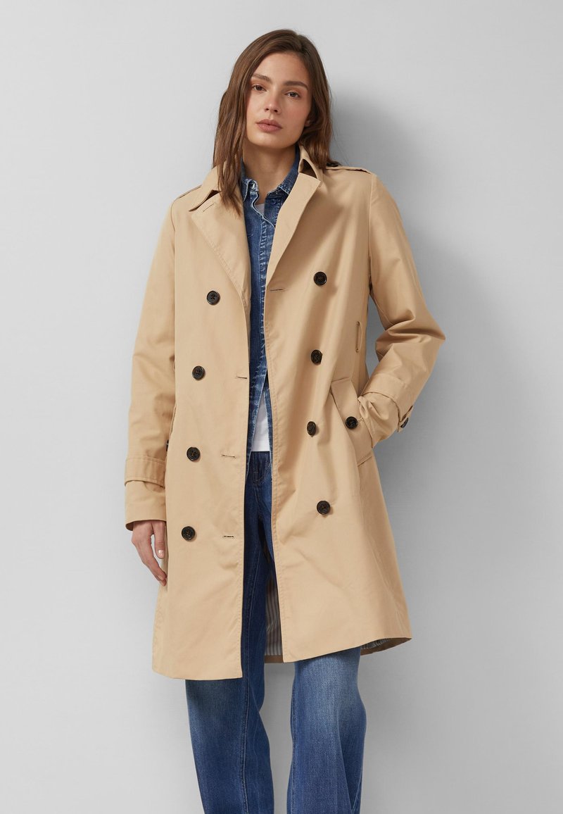 Frau trägt einen beigefarbenen, doppelt geknöpften Trenchcoat über einer Jeansjacke und blauen Jeans, steht mit einer Hand in der Tasche vor einer grauen Wand.