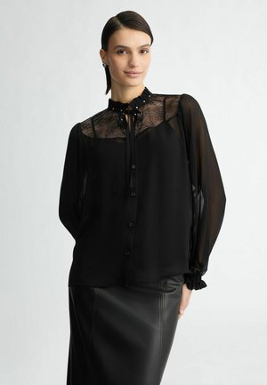 Blusa nera con corpetto in pizzo, colletto a volant e chiusura con bottoni, dotata di maniche lunghe e una texture trasparente, abbinata a una gonna in pelle nera.