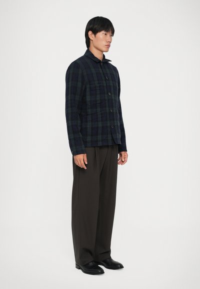 Giacca a quadri in navy scuro e verde, realizzata in tessuto morbido, con chiusura a bottoni, colletto e due tasche sul petto. Abbinata a pantaloni scuri.