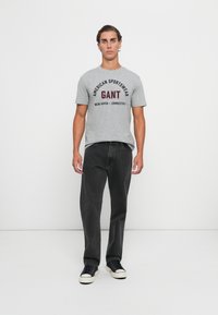 Mann står, iført grå GANT T-skjorte med tekst og svarte jeans, mot en ensfarget hvit bakgrunn.