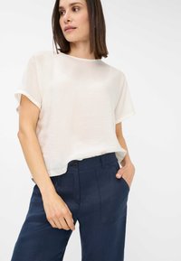 Blusa tagliata, leggera, di colore bianco sporco con maniche arrotolate, abbinata a pantaloni blu navy, sartoriali, con fronte piatto e tasche laterali.