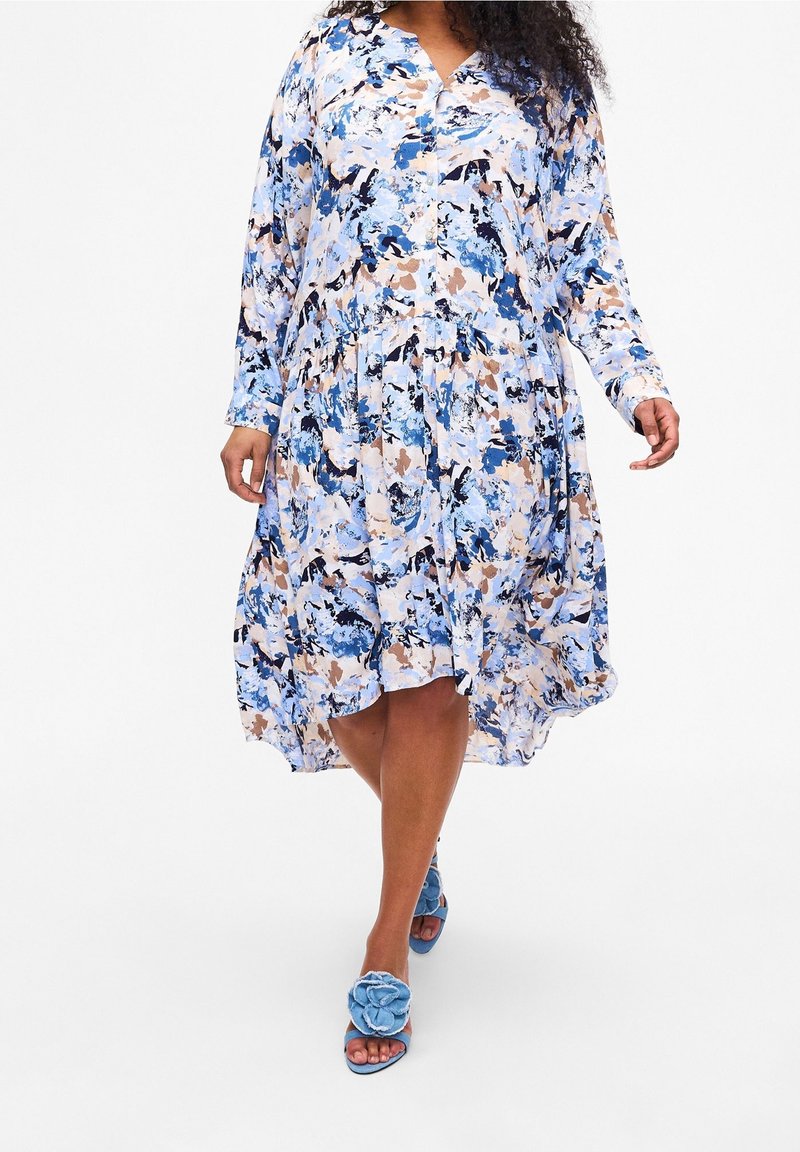 Modré květinové šaty s dlouhými rukávy, v-výstřihem a nařaseným pasem, s volným lemem. Spárovány s modrými botami s květinovými akcenty.
