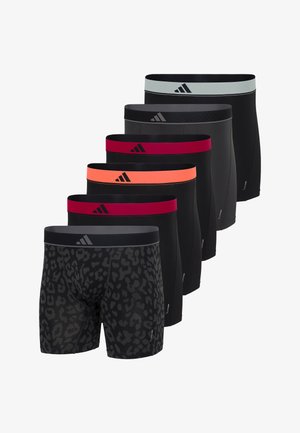 Muskuløs mand stående barfodet iført sorte boksershorts med rød linning og Adidas-logo, kigger til siden mod en hvid baggrund.