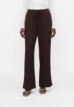 Persoană care poartă pantaloni largi maro închis cu șnur în talie, un top alb cu mâneci lungi și sandale bej cu toc și vârf decupat.