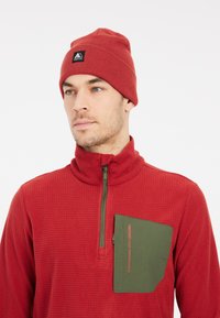 Rode gebreide pullover met een halflange ritskraag, voorzien van een groene borstzak en een contrasterend logo. Een bijpassende rode gebreide beanie.
