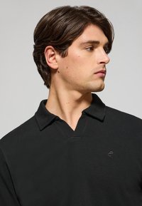 Svart polo-skurt med ett texturerat tyg, med en modern krage och en liten logotyp på bröstet. Ren och minimalistisk stil.