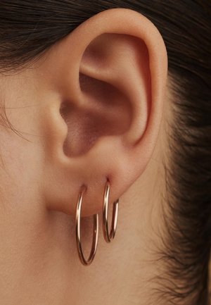 Boucles d'oreilles en métal rose gold, présentant deux cercles liés d'une texture lisse, suspendus en dessous d'une oreille humaine pour un détail en gros plan.