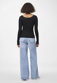 Person med lockigt hår som bär en svart långärmad tröja och ljusblå vida jeans, stående med ryggen mot en enfärgad bakgrund.