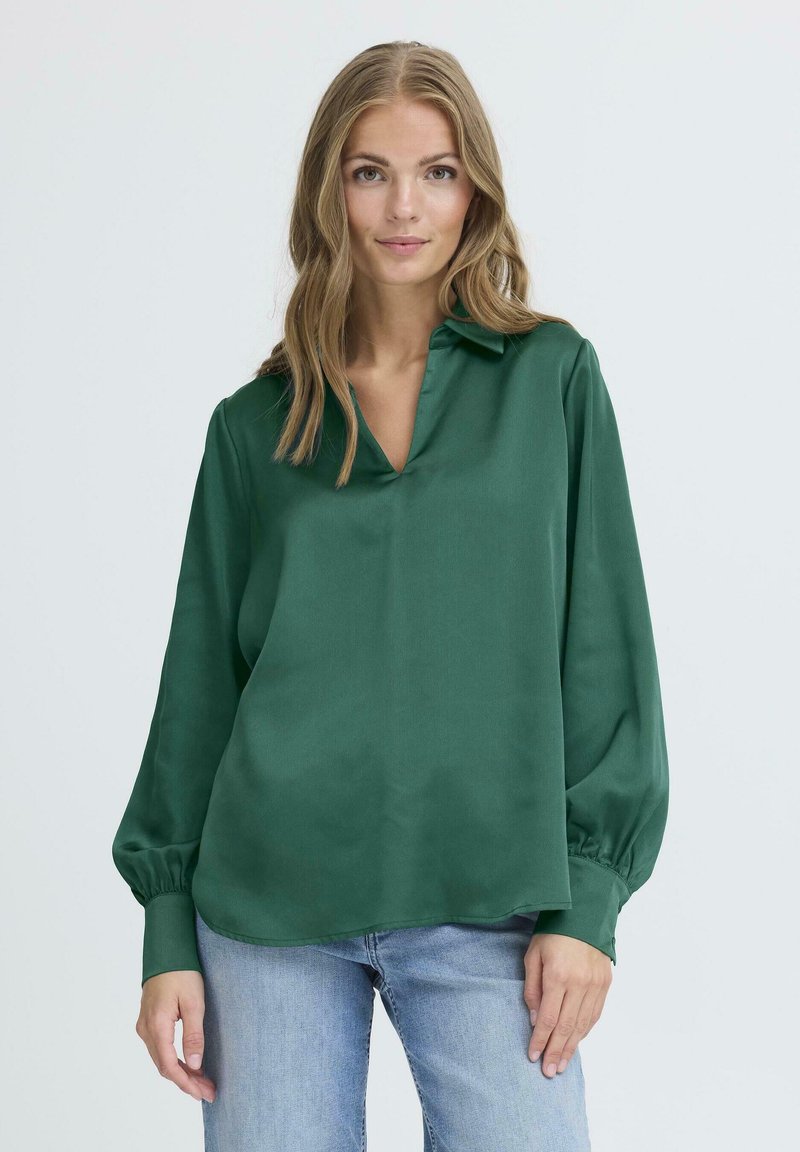 Blusa di seta verde con scollo a V, maniche lunghe a palloncino e polsini con bottoni, abbinata a jeans in denim blu chiaro. Tessuto liscio e leggero.