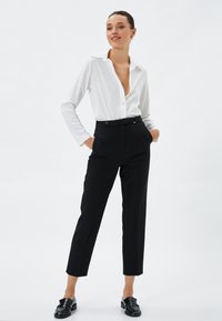 Hvid langærmet skjorte med dyb V-udskæring, parret med sorte højtaljede bukser. Sorte loafers fuldender outfittet.