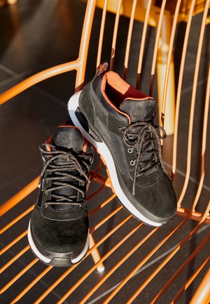 Paire de chaussures de sport noires avec semelles blanches et intérieur orange, posées côte à côte sur une chaise en métal orange en plein soleil.