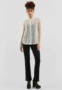 Transparante kantblouse in ivoorkleur met een knoopsluiting en kraag, heeft lange mouwen over een strakke zwarte outfit en dikke zwarte plateauschoenen.