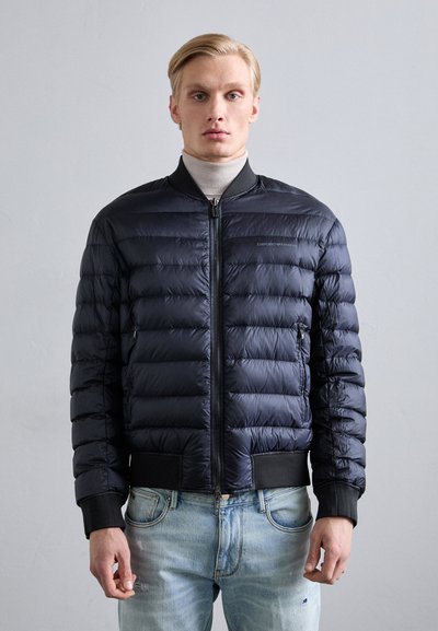 Marinblå puffjacka med horisontellt quiltat mönster, svart ribbad krage och manschetter, dragkedjestängning och logotypdetalj på bröstet. Bärs över ljusa jeans.