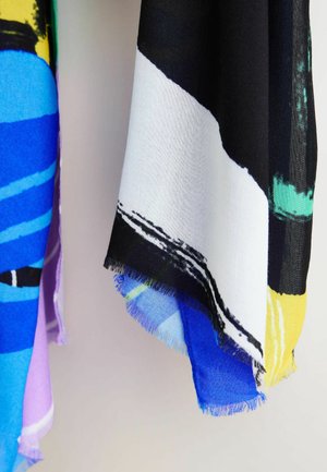 Kosički tkanine z abstraktnimi vzorci v črni, beli, modri, rumeni, vijolični in zeleni barvi, ki visijo na enobarvni svetli podlagi.