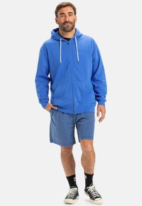 Blue zip-up hoodie with drawstrings, soft fabric, kangaroo pocket; paired with light blue shorts and black sneakers.