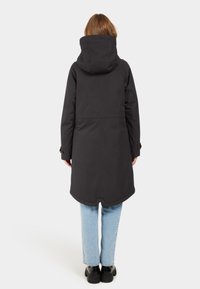 Langer schwarzer Mantel mit Kapuze, aus glattem, wasserabweisendem Stoff. Hat einen verlängerten Rücken und geknöpfte Manschetten. Wird mit hellen Jeans kombiniert.