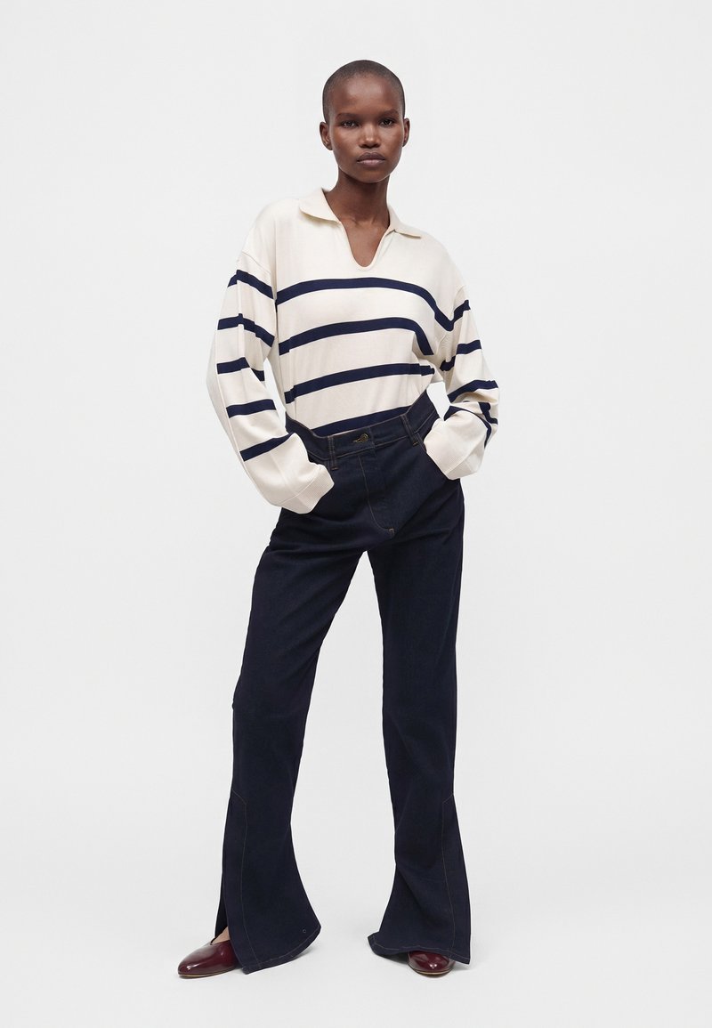 Personne debout portant un pull blanc avec de larges rayures bleu marine, un pantalon noir évasé taille haute et des chaussures rouge foncé, les mains dans les poches.