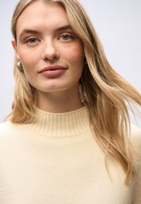 Pull en tricot beige avec col montant, texture douce, coupe décontractée et manches longues. Le modèle a les cheveux blonds et se trouve devant un arrière-plan neutre.