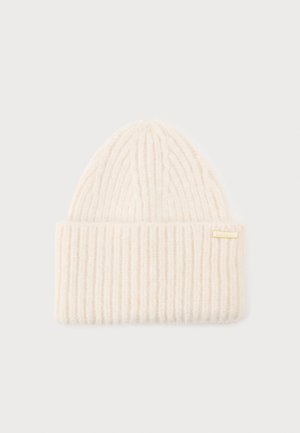 Beige knitted beanie hat with a ribbed texture, featuring a cuff and a small gold metal logo tag. Soft material and elongated top shape.