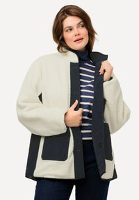 Giacca in pile bianca con dettagli e bottoni blu navy. Presenta un taglio oversize, un design trapuntato e grandi tasche frontali. Sotto è indossato un dolcevita a righe.