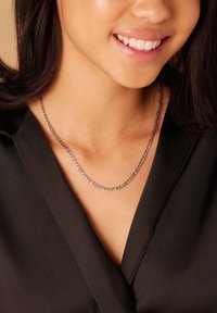 Jeune personne souriante, portant un collier en chaîne argentée et un haut noir à décolleté en V, avec des cheveux foncés partiellement visibles.