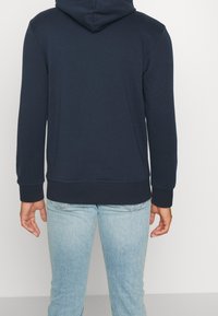 Sudadera de color azul marino hecha de una tela suave, con un bolsillo tipo canguro, puños y dobladillo acanalados; cuenta con una capucha ajustable y un corte holgado.