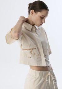 Kurzes, tailliertes beige Oberteil mit aufwendiger floraler Stickerei an den Ärmeln. Der Stoff hat eine glatte, leichte Textur.