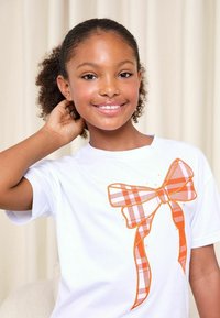 Maglietta bianca con grande fiocco a quadri arancioni e bianchi stampato sul davanti. Tessuto liscio con scollo rotondo e maniche corte.