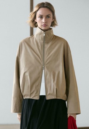 Kvinde med kort hår iført en beige lynlåstrøje, hvid top og sort nederdel, der holder et rødt klæde op mod en ensfarvet baggrund.