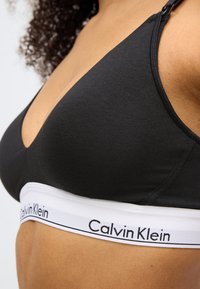 Crni trokutasti grudnjak od mekanog materijala s bijelom elastičnom trakom na kojoj je logo "Calvin Klein". Podesive crne naramenice za bolje prilagođavanje.
