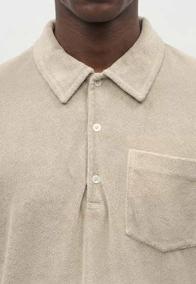 Κοντινό πλάνο προσώπου που φοράει μπεζ υφασμάτινη πόλο μπλούζα με τρία κουμπιά και τσέπη στο στήθος.