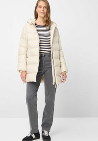 Steppjacke in Cremeton mit Kapuze, mit Steppmuster, Reißverschluss und Seitentaschen; kombiniert mit grauen Jeans und Turnschuhen.