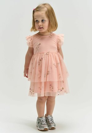 Niña pequeña con cabello rubio, vestida con un vestido rosa de tul con patrones de estrellas y zapatillas plateadas, de pie sobre un fondo liso.