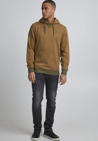 Felpa marrone con tasca a marsupio e dettagli verdi, abbinata a jeans scuri e sneakers nere. Tessuto liscio, vestibilità casual.