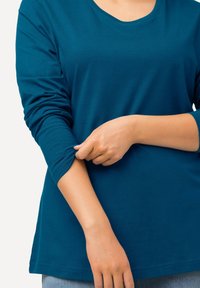 Tyrkysové tričko s dlouhým rukávem z měkké látky, s kulatým výstřihem a mírně ohrnutými rukávy, spojené s džínovými kalhotami.