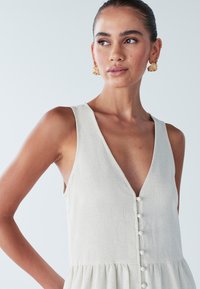 Jasno-beżowa lniana sukienka z głębokim dekoltem w kształcie V, bez rękawów oraz z guziczkami z przodu. Spódnica delikatnie poszerza się na dole.