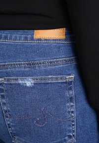 Jeans de mezclilla azul oscuro con una etiqueta de cuero con logotipo en relieve, costuras visibles y un detalle desgastado en el bolsillo trasero.