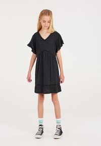 Dunkelgraues V-Ausschnittkleid mit kurzen Rüschensäumen, gerafftem Taillenbereich und abgestuftem Saum, getragen mit schwarzen High-Top-Schuhen und grün gestreiften Socken.